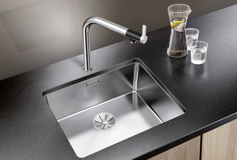 Na obrazie widoczny jest srebrny zlewozmywak jednokomorowy wbudowany w ciemny blat kuchenny, z nowoczesną baterią. Obok zlewozmywaka znajduje się karafka z wodą i dwie szklanki.