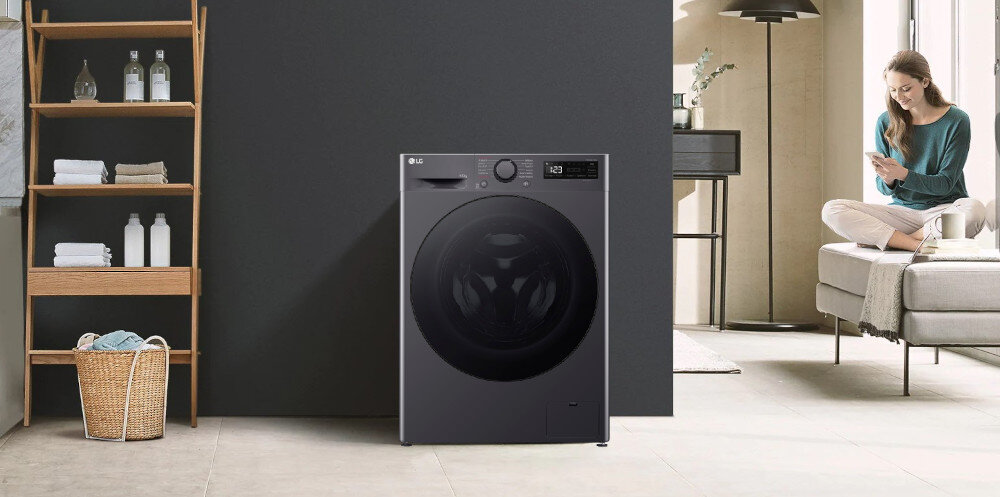 Pralko-suszarka w kolorze ciemnym stoi na tle ciemnej ściany, obok drewnianego regału z ręcznikami i butelkami. Na panelu widoczne jest logo LG oraz wyświetlacz z przyciskami.