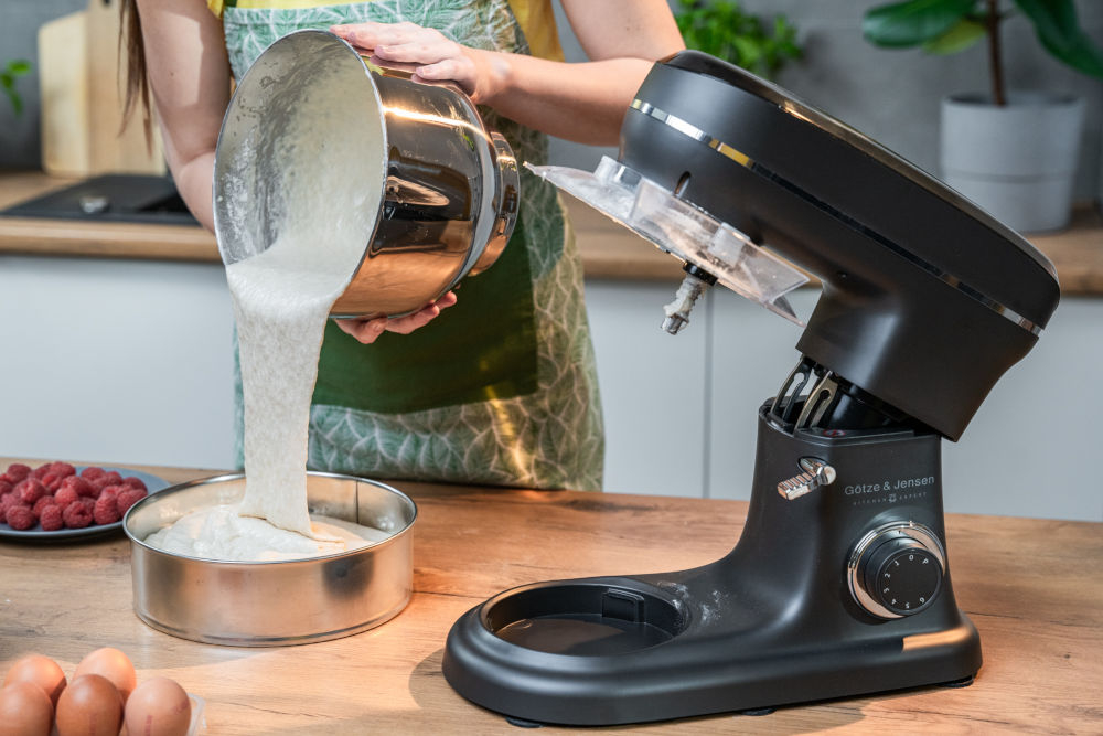 Osoba wlewa ciasto z metalowej miski do okrągłej formy na blacie kuchennym. Obok znajduje się czarny robot kuchenny Götze & Jensen z widocznym logo.