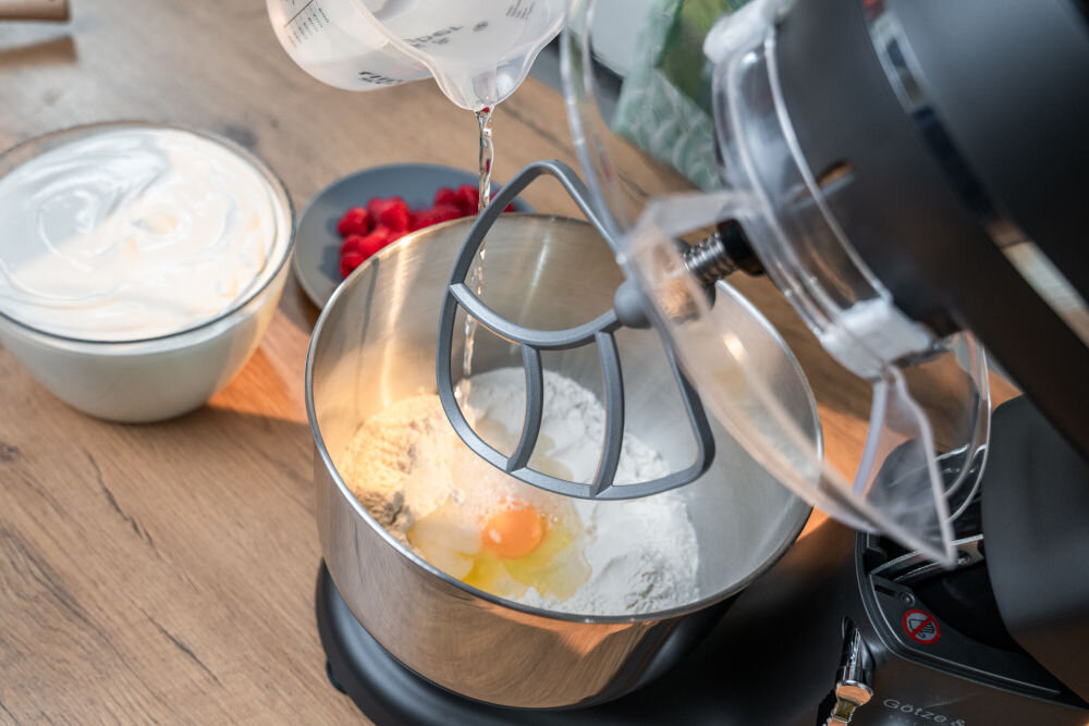 Robot kuchenny planetarny miesza składniki w metalowej misie, w której znajdują się mąka i jajko. Obok stoi miska z ubitym kremem i talerzyk z malinami.