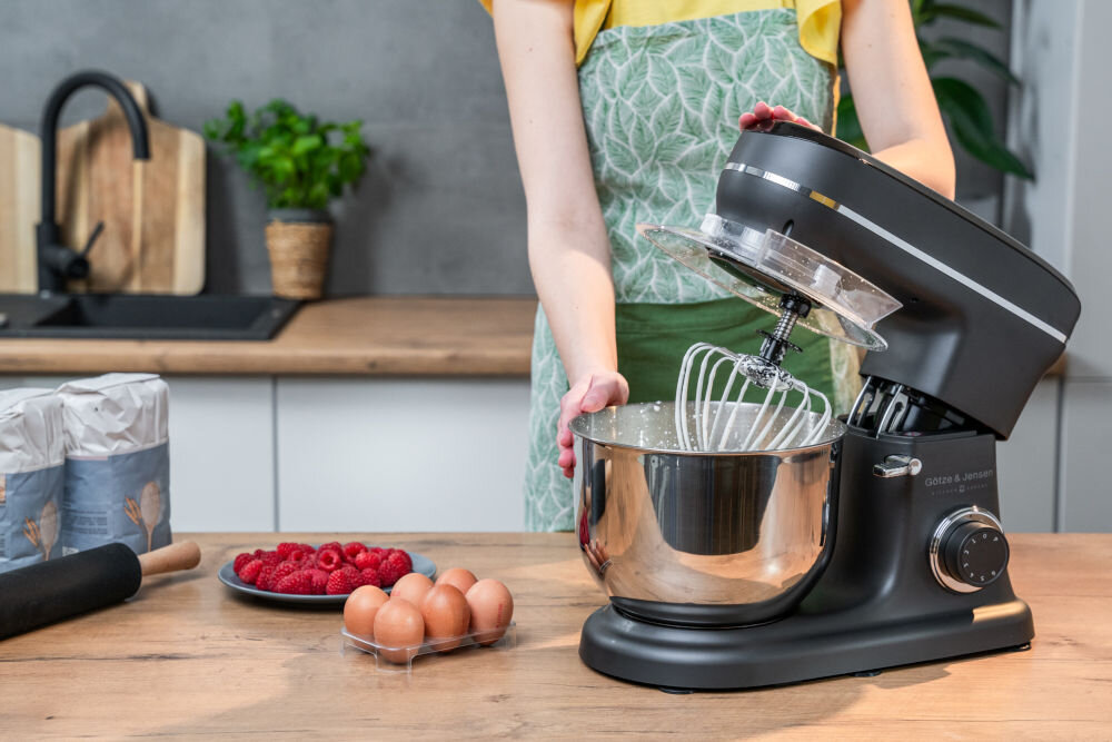 Na blacie kuchennym znajduje się robot kuchenny planetarny GÖTZE & JENSEN SB400K z misą i trzepaczką. Obok leżą jajka, maliny i opakowania z mąką.