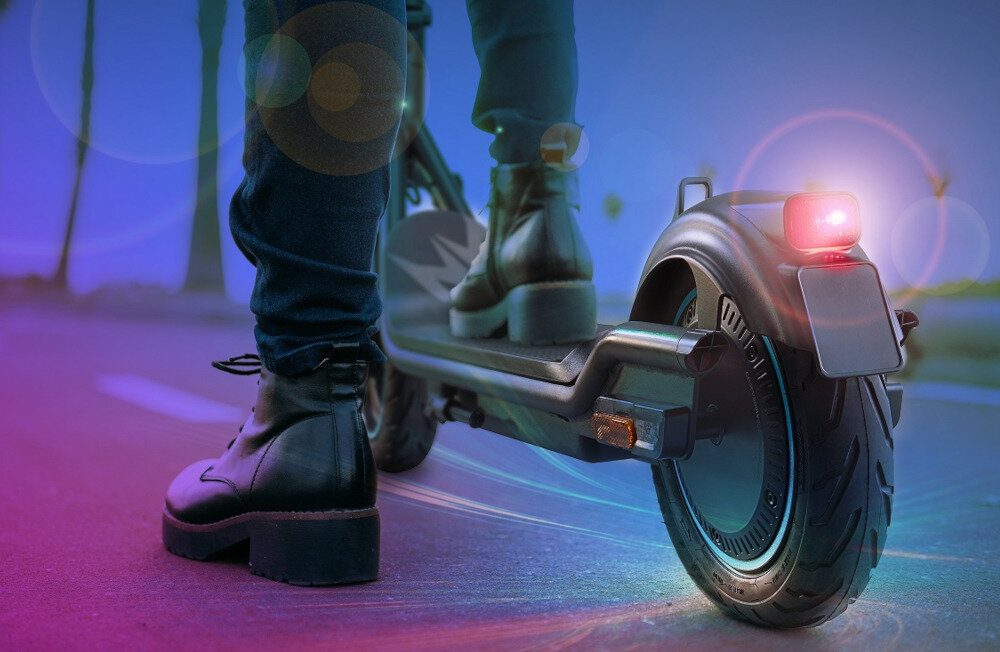 Osoba stoi na elektrycznej hulajnodze z dużym tylnym kołem i świecącym czerwonym światłem. Tło jest rozmyte, z kolorowymi efektami świetlnymi.
