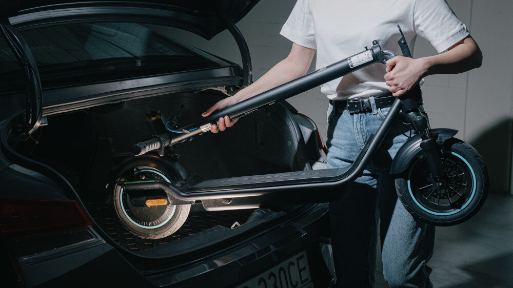 Osoba trzyma czarną hulajnogę elektryczną, wyciągając ją z bagażnika samochodu. Hulajnoga ma duże koła i widoczne elementy amortyzacji.