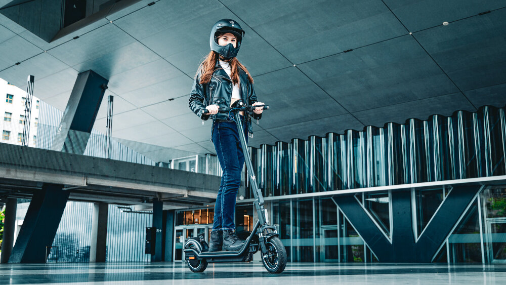 Osoba stoi na czarnej hulajnodze elektrycznej w nowoczesnym, miejskim otoczeniu. Hulajnoga ma duże koła i solidną konstrukcję.