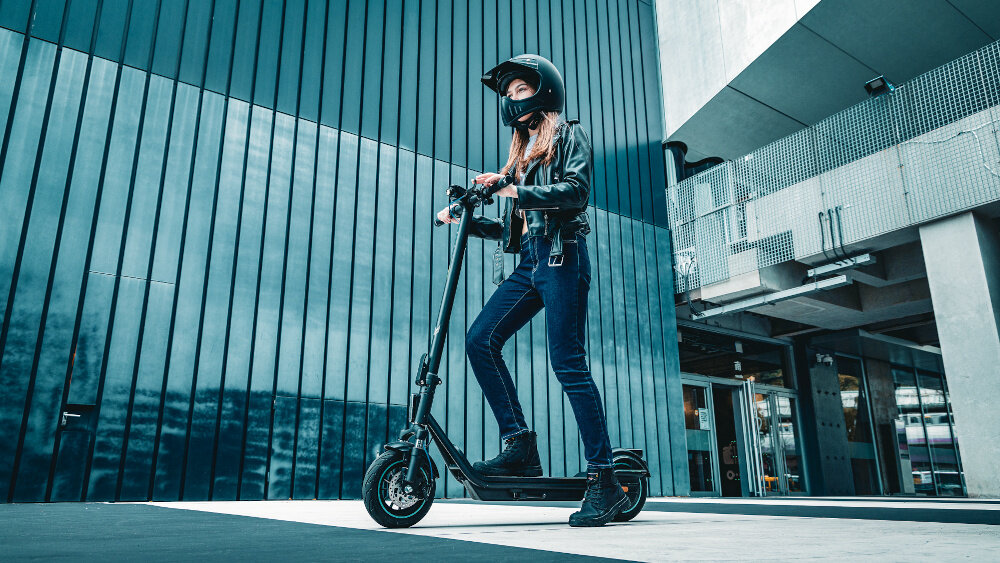 Osoba w kasku stoi na czarnej hulajnodze elektrycznej przed nowoczesnym budynkiem z metalowymi panelami. Hulajnoga ma duże koła i solidną konstrukcję.