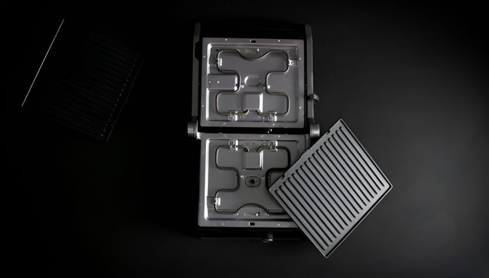 Obraz przedstawia otwarty grill elektryczny z widocznymi elementami grzewczymi i jedną wyjętą kratką grillową. Tło jest ciemne, co podkreśla metaliczne elementy urządzenia.