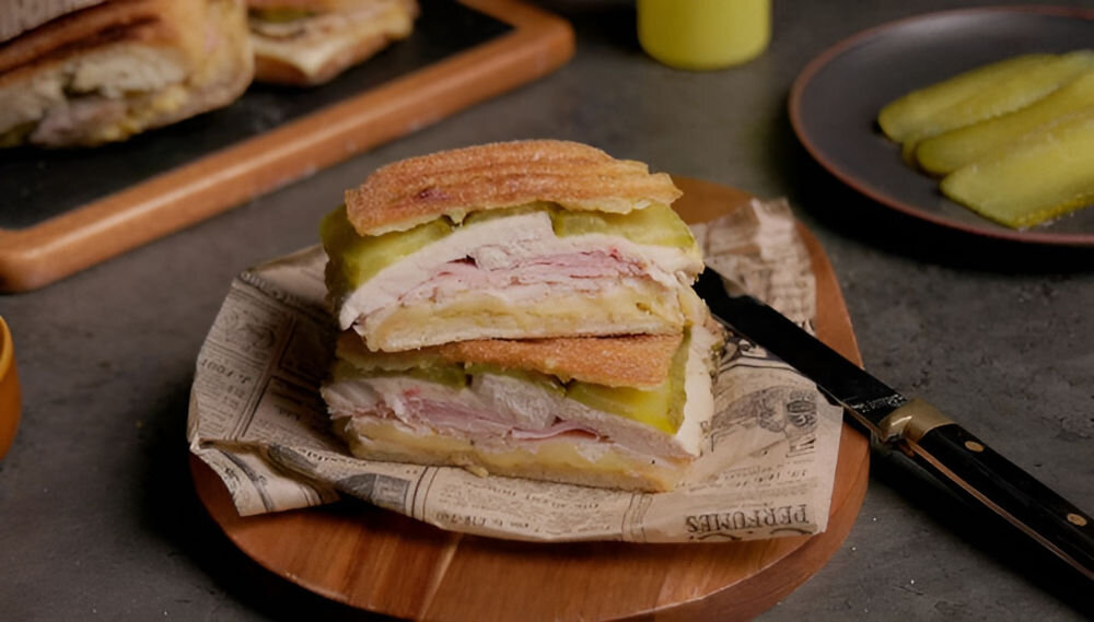 Na drewnianej desce leży kanapka złożona z kilku warstw mięsa, sera i ogórka, owinięta w papier z nadrukiem. Obok znajduje się nóż, a w tle widoczny jest talerz z plasterkami ogórka.