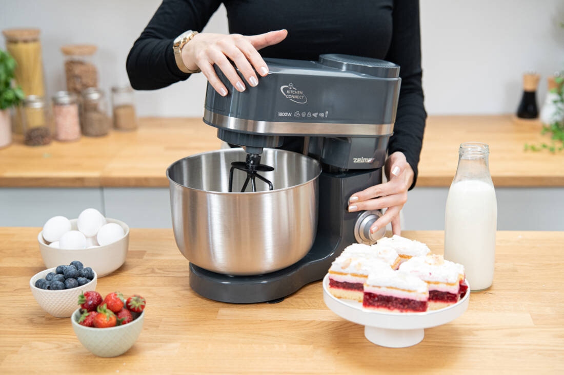 Na drewnianym blacie kuchennym znajduje się robot kuchenny z metalową misą, obok którego leży butelka mleka i talerz z ciastem. W tle widoczne są miski z owocami i jajkami.