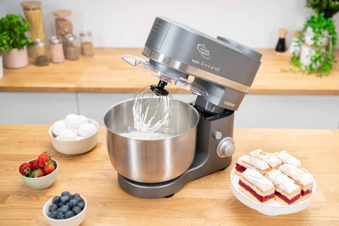 Na drewnianym blacie kuchennym stoi robot kuchenny planetarny z metalową misą, w której znajduje się ubijana masa. Obok robota znajdują się miski z truskawkami, borówkami, jajkami oraz talerz z ciastem.