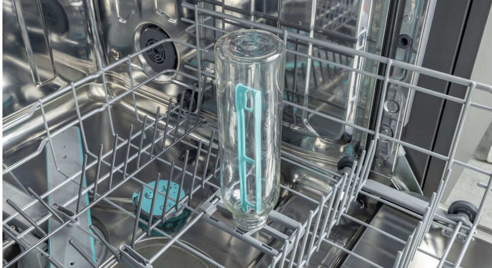 Wnętrze zmywarki z widocznym szklanym naczyniem umieszczonym w górnym koszu. W tle widać metalowe elementy konstrukcji zmywarki oraz niebieskie detale plastikowe.