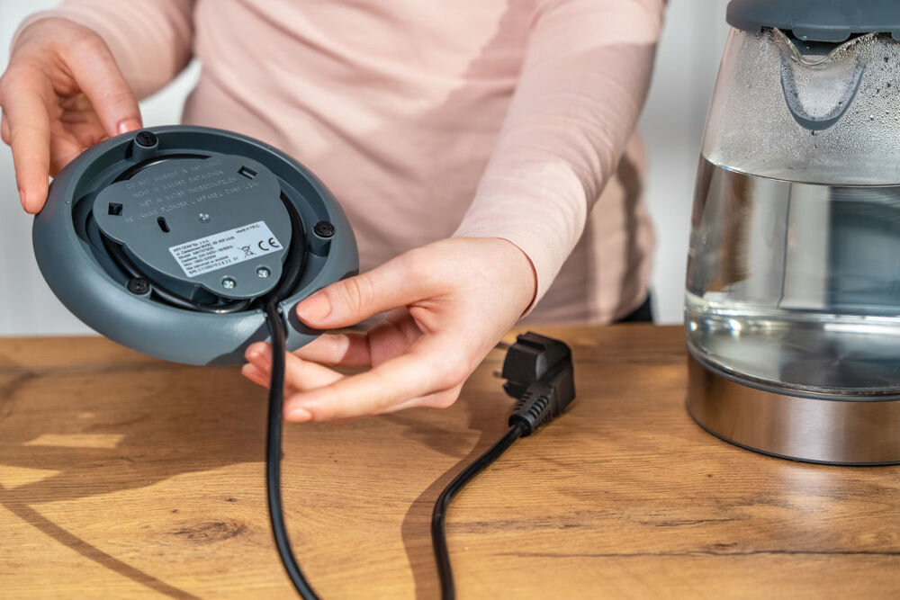 Osoba trzyma podstawę czajnika elektrycznego z widocznym kablem zasilającym. Obok znajduje się szklany czajnik z wodą na drewnianym blacie.