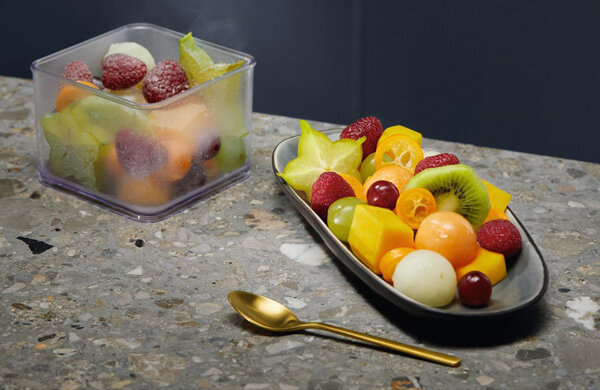 Na blacie z kamienia znajduje się prostokątne naczynie z przezroczystego tworzywa, wypełnione mrożonymi owocami. Obok leży owalny talerz z różnorodnymi świeżymi owocami oraz złota łyżeczka.