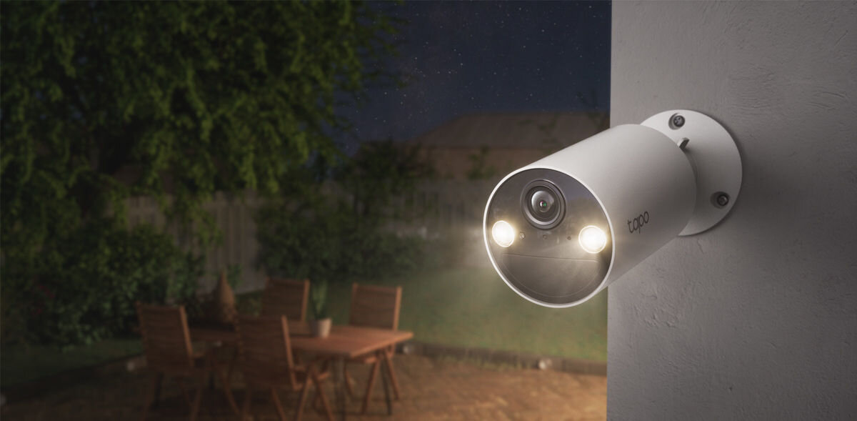 Na obrazie widoczna jest zewnętrzna kamera zamontowana na ścianie, z dwoma świecącymi diodami. W tle znajduje się ogród z drewnianym stołem i krzesłami.