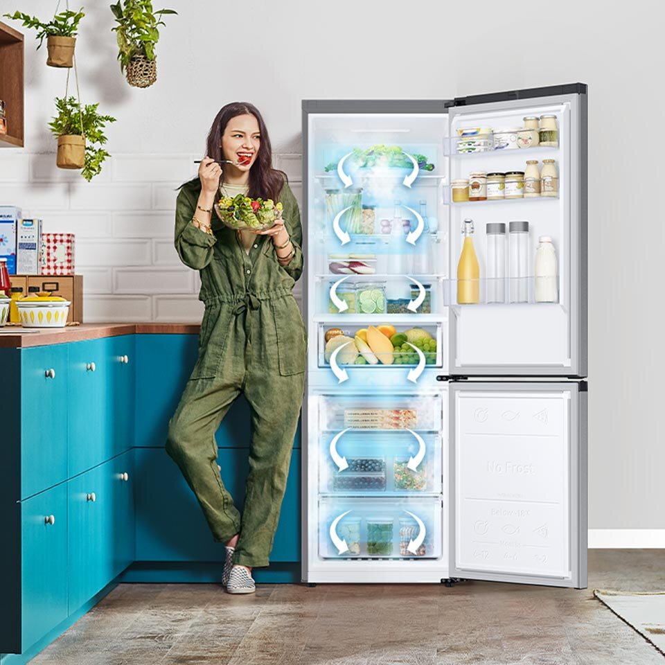 Kobieta stoi obok otwartej lodówki w kuchni, trzymając miskę sałatki. W lodówce widoczne są różne produkty spożywcze, a na drzwiach znajdują się butelki i słoiki.