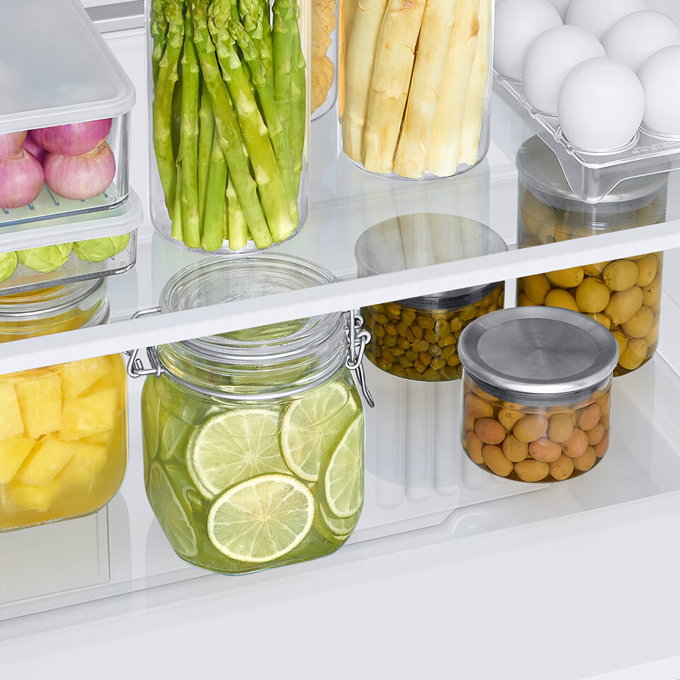 Wnętrze lodówki z różnorodnymi produktami spożywczymi w słoikach i pojemnikach, w tym szparagi, cytryny, jajka i inne warzywa. Produkty są starannie ułożone na szklanych półkach.