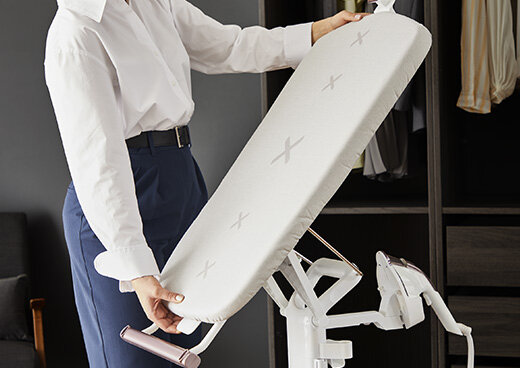 Osoba trzyma deskę do prasowania, która jest częścią systemu do prasowania z białym pokrowcem i wzorem w kształcie litery X. W tle widoczna jest szafa z wiszącymi ubraniami.