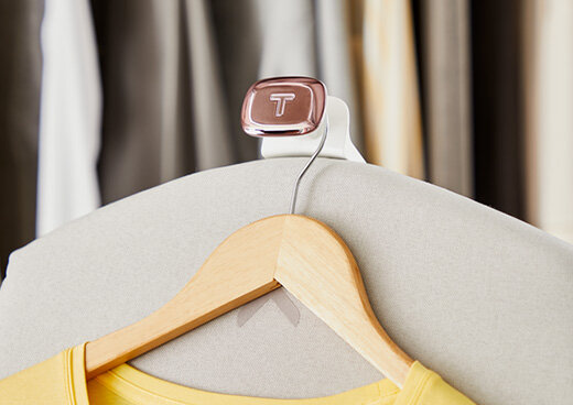 Na obrazie widoczny jest wieszak z drewnianą poprzeczką, na którym zawieszona jest żółta koszulka. W tle znajdują się rozmyte elementy garderoby, a na wieszaku widnieje logo \'T\'.