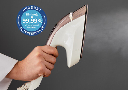 Osoba trzyma żelazko, z którego wydobywa się para. Obok widoczny jest niebieski okrągły znak z napisem: \'PRODUKT DEZYNFEKUJĄCY Eliminuje do 99,99% wirusów, bakterii i zarazków\'.