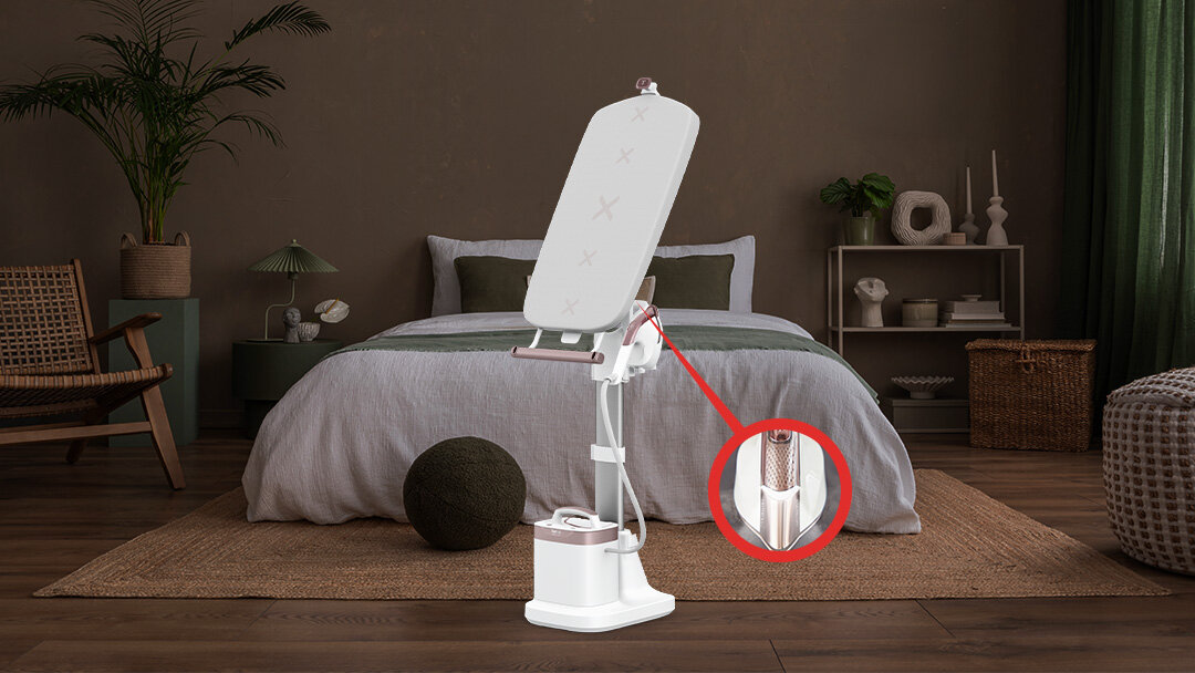 Na obrazie znajduje się system do prasowania z deską i stojakiem, umieszczony w sypialni z łóżkiem i roślinami w tle. W prawym dolnym rogu widoczny jest zbliżony fragment urządzenia z oświetleniem LED.