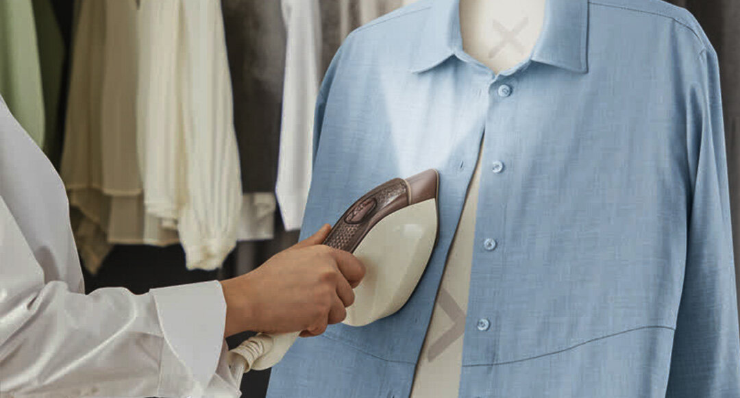 Osoba prasuje niebieską koszulę za pomocą urządzenia do prasowania z oświetleniem LED. W tle widoczne są inne ubrania na wieszakach.