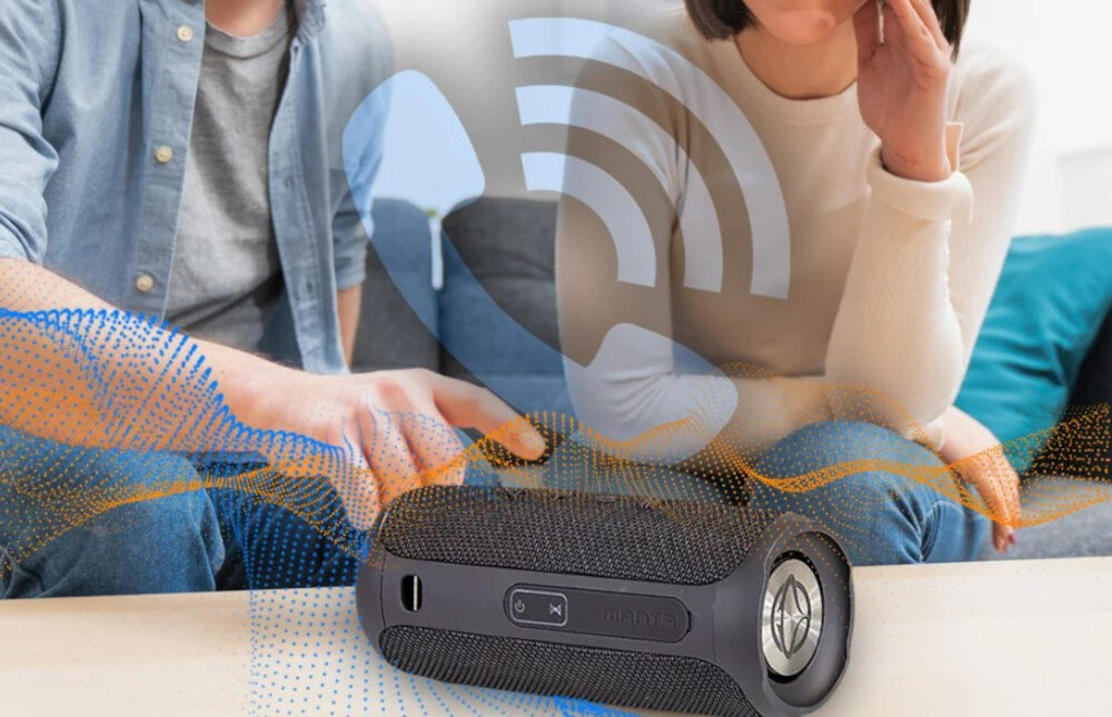 Na stole leży czarny głośnik mobilny z widocznym logo \'manta\' i przyciskami. W tle widać dwie osoby siedzące na kanapie oraz graficzne elementy symbolizujące dźwięk.