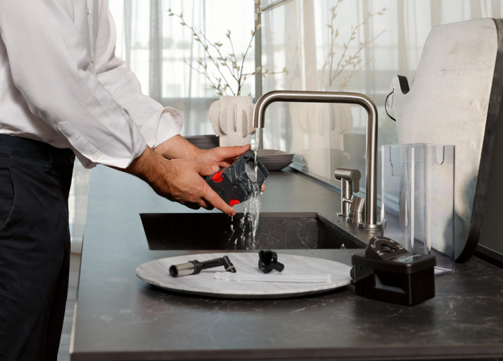 Osoba myje element ekspresu do kawy pod bieżącą wodą w zlewie. Na blacie obok znajdują się inne części ekspresu oraz taca.