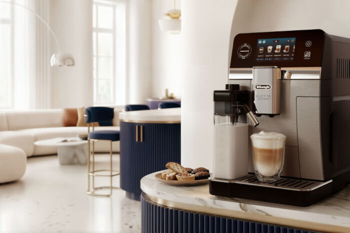 Na blacie kuchennym stoi ekspres do kawy z logo \'DeLonghi\', obok którego znajduje się szklanka z kawą i spieniacz do mleka. W tle widoczna jest elegancka kuchnia z niebieskimi krzesłami i jasnymi ścianami.