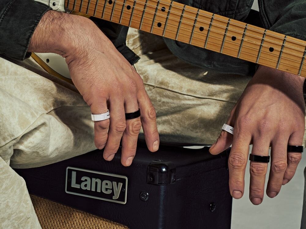 Osoba trzyma gitarę i ma na palcach kilka pierścieni w kolorach białym i czarnym. Na wzmacniaczu widoczne jest logo \'Laney\'.