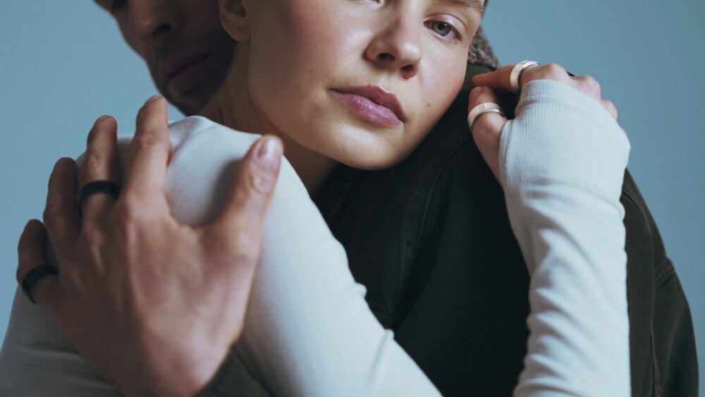 Dwie osoby obejmują się, obie mają na palcach białe i czarne pierścienie. Tło jest jednolite, w odcieniu niebieskim.