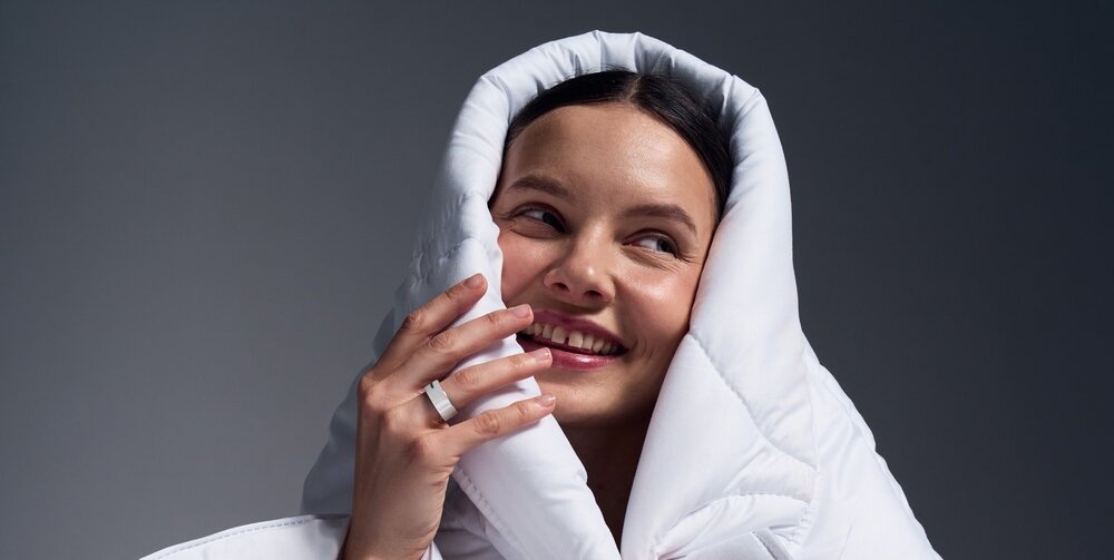 Osoba owinięta w biały materiał, uśmiecha się, nosząc biały pierścień na palcu. Tło jest jednolite i ciemne.