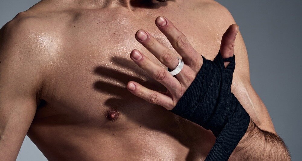 Osoba z owiniętą dłonią w czarny bandaż prezentuje biały pierścień na palcu. Tło jest neutralne, a skóra osoby jest lekko błyszcząca.