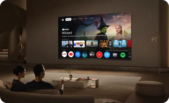 W przestronnym salonie na ścianie wisi duży telewizor wyświetlający interfejs Google TV z różnymi aplikacjami i filmem \'Wicked\'. Przed telewizorem siedzą dwie osoby na kanapie, a na stoliku przed nimi stoi wazon z rośliną.