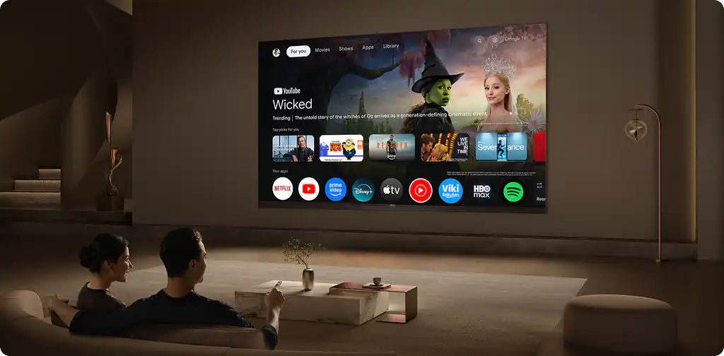Na ścianie wisi duży telewizor wyświetlający interfejs Google TV z aplikacjami takimi jak YouTube, Netflix i Spotify. Przed telewizorem siedzą dwie osoby na sofie, a na stoliku przed nimi stoi mała roślina.