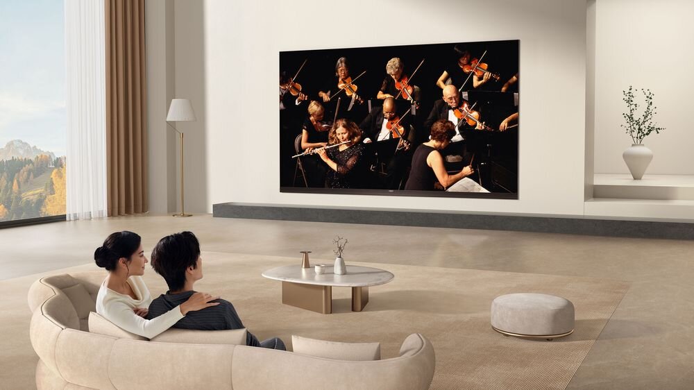 W przestronnym salonie na ścianie wisi duży telewizor, na którym wyświetlany jest koncert orkiestry. Na kanapie siedzą dwie osoby oglądające program.