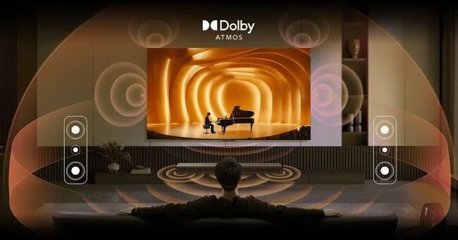 Osoba siedzi na kanapie przed dużym telewizorem, na którym wyświetlany jest obraz pianisty w pomarańczowym tunelu. Na górze ekranu widnieje logo \'Dolby Atmos\'.