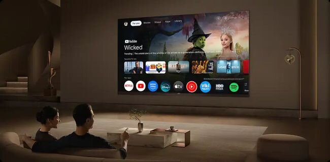 Na ścianie wisi duży telewizor wyświetlający interfejs Google TV z aplikacjami takimi jak YouTube, Netflix i Spotify. Przed telewizorem siedzą dwie osoby na sofie, a na stoliku przed nimi stoi mała roślina.