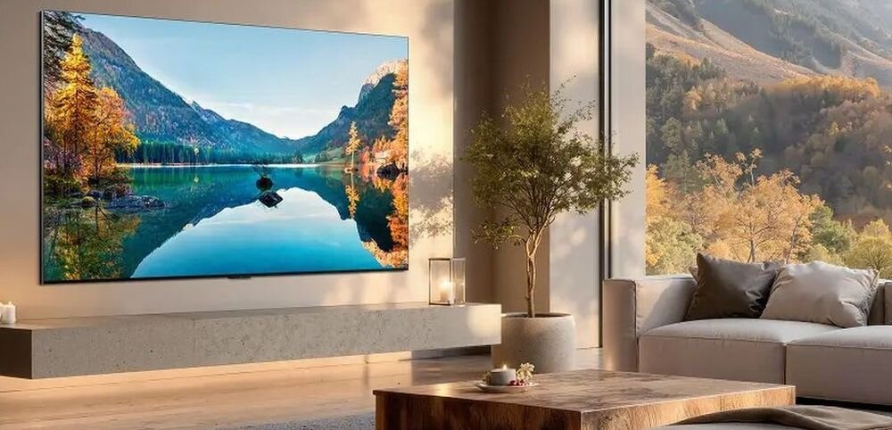 W nowoczesnym salonie na ścianie wisi duży telewizor wyświetlający krajobraz z górami i jeziorem. W pomieszczeniu znajduje się także sofa, stolik i roślina w doniczce.
