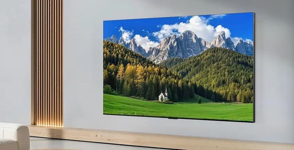 Na ścianie wisi duży telewizor wyświetlający obraz górskiego krajobrazu z lasem i niebem pełnym chmur. W tle widoczne są drewniane panele i jasna ściana.