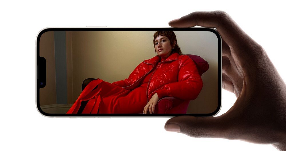 Osoba ubrana w czerwony płaszcz siedzi na krześle, widoczna na ekranie smartfona trzymanego w dłoni. Smartfon ma cienkie ramki i zaokrąglone rogi.