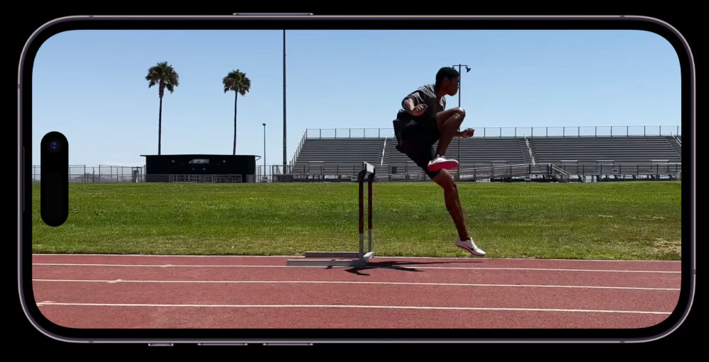 Obraz przedstawia smartfon z wyświetlaczem, na którym widać osobę przeskakującą przez przeszkodę na bieżni. W tle znajdują się trybuny i dwie palmy.