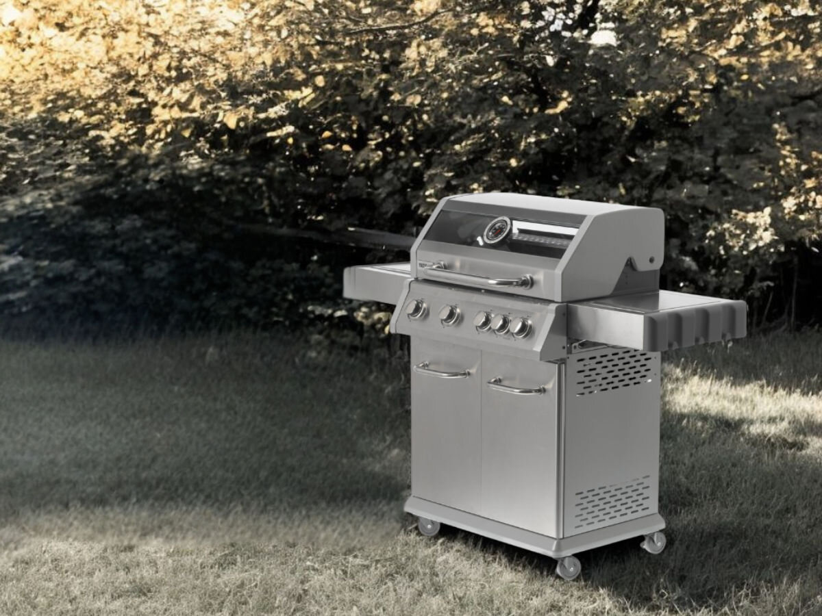 Na trawniku stoi duży, metalowy grill gazowy z pokrywą i kilkoma pokrętłami. W tle widoczne są drzewa i krzewy.