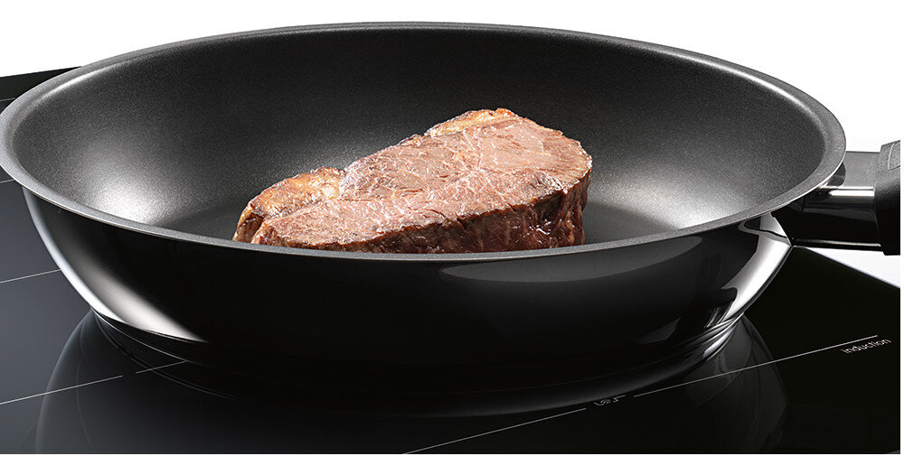 Na płycie indukcyjnej znajduje się patelnia z kawałkiem mięsa. Powierzchnia płyty jest czarna z delikatnymi liniami i oznaczeniami stref grzewczych.