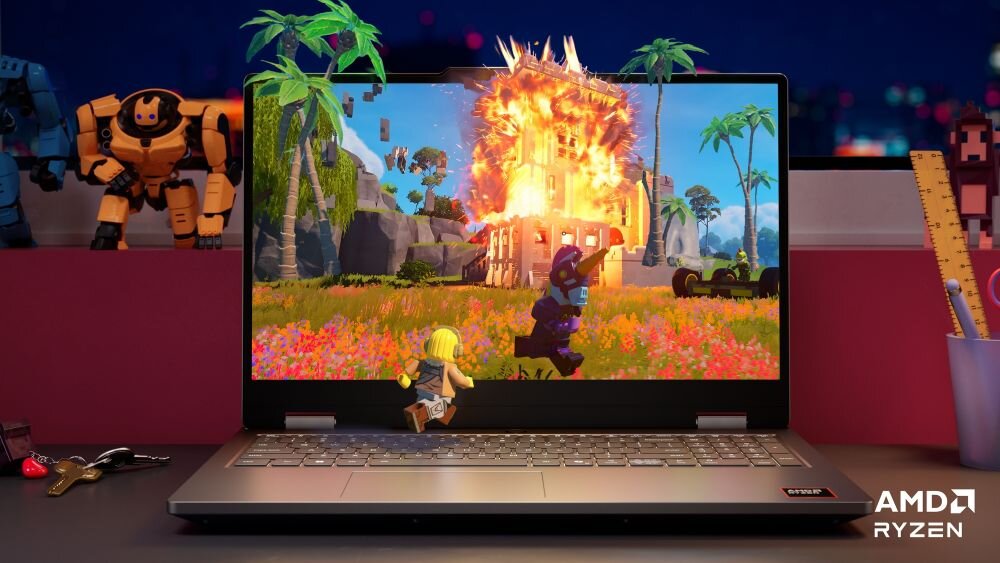 Laptop z wyświetlaczem pokazującym grę z postacią biegnącą przed płonącym budynkiem. Na biurku obok laptopa znajdują się figurki robotów i linijka, a w rogu ekranu widoczne jest logo AMD Ryzen.