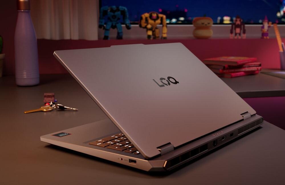Na biurku znajduje się otwarty laptop z widocznym logo \'LOQ\' na pokrywie. W tle widać figurki i książki na parapecie.