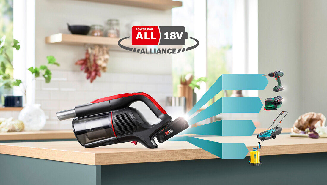 Na blacie kuchennym znajduje się odkurzacz ręczny w kolorze czarno-czerwonym. W tle widoczne jest logo \'POWER FOR ALL 18V ALLIANCE\' oraz grafika przedstawiająca różne narzędzia zasilane tym samym systemem.