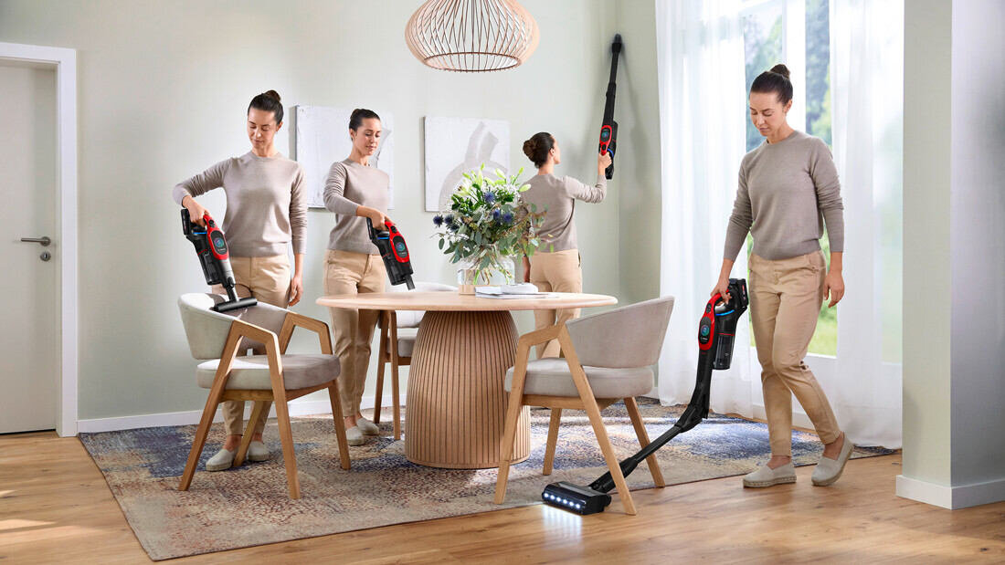 Cztery kobiety w beżowych ubraniach używają odkurzacza w jasnym, nowoczesnym salonie z drewnianą podłogą i dużym stołem. Odkurzacz ma czarno-czerwony kolor i jest używany do czyszczenia różnych powierzchni, w tym podłogi i mebli.