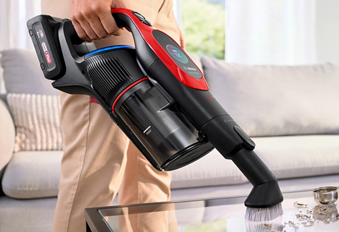 Osoba trzyma odkurzacz ręczny z widocznym logo \'BOSCH\', używając go do czyszczenia szklanej powierzchni. W tle znajduje się jasna sofa i zasłony.