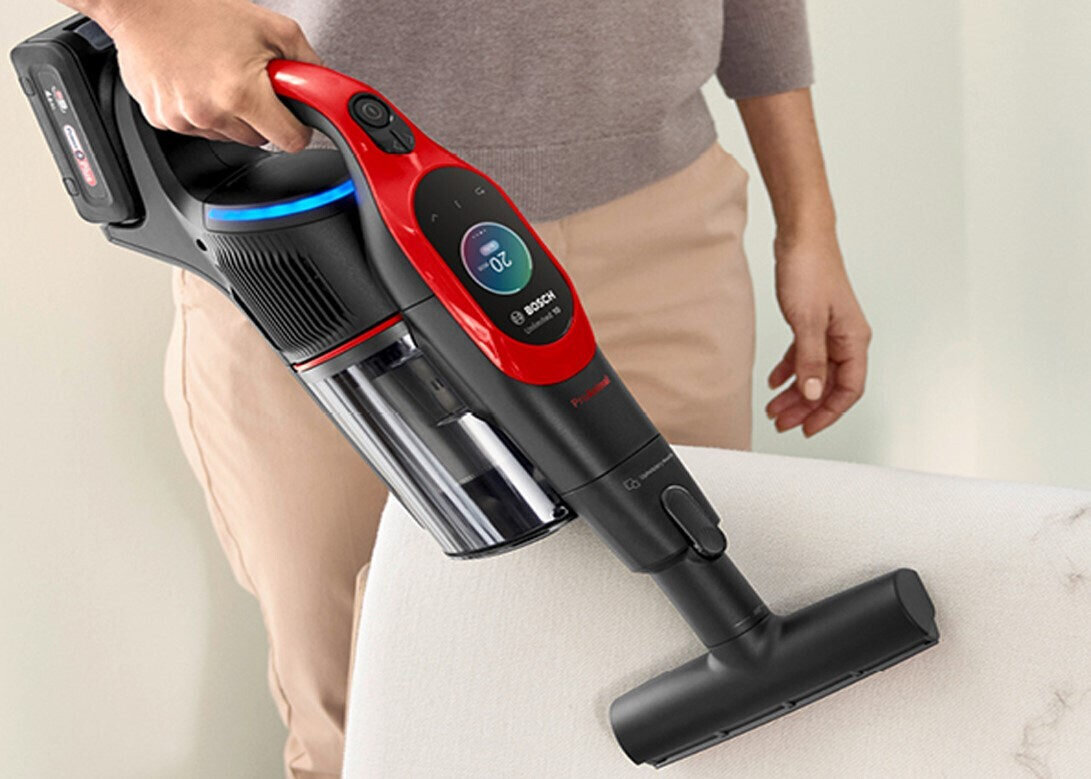 Osoba trzyma odkurzacz ręczny marki BOSCH z widocznym logo i wyświetlaczem pokazującym \'20%\'. Odkurzacz jest używany do czyszczenia jasnej powierzchni.