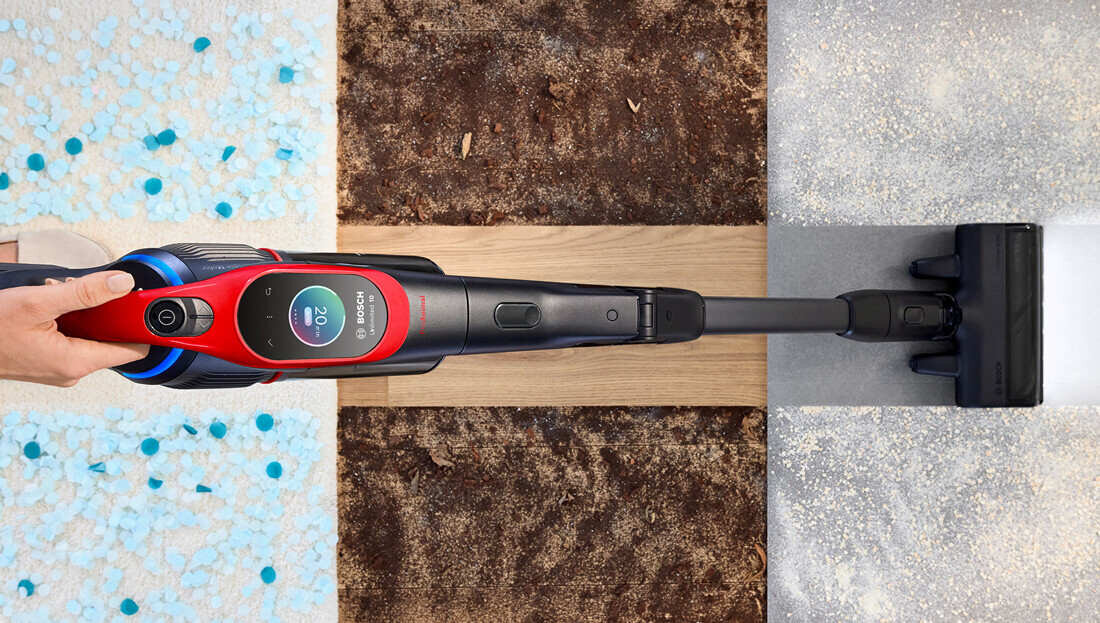 Osoba trzyma odkurzacz z widocznym logo \'BOSCH\' na wyświetlaczu, który pokazuje liczbę \'20\'. Odkurzacz jest używany na różnych powierzchniach: dywanie, drewnie i płytkach.