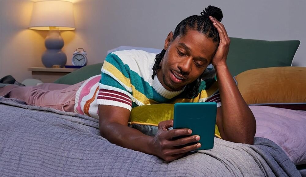 Osoba leży na łóżku, trzymając w ręku czytnik e-booków. W tle widoczna jest lampa i zegar na stoliku nocnym.
